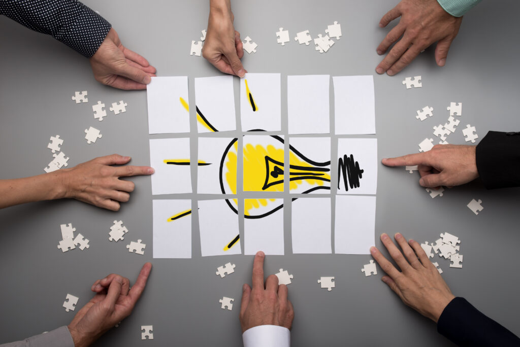 Multiple hands collaborating to assemble a lightbulb puzzle, symbolizing teamwork, problem-solving, and creativity as essential real-life skills for generating new ideas.