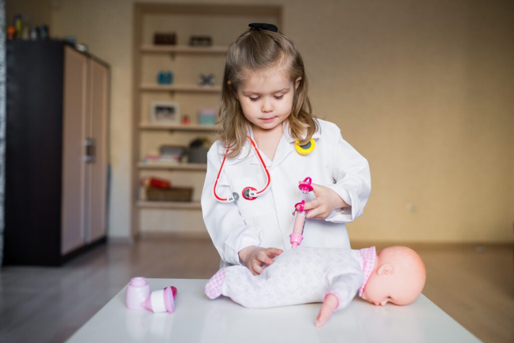 Young child dressed as a doctor, giving a toy injection to a doll, symbolizing the importance to stop forcing kids to be doctors or engineers and let them explore freely.