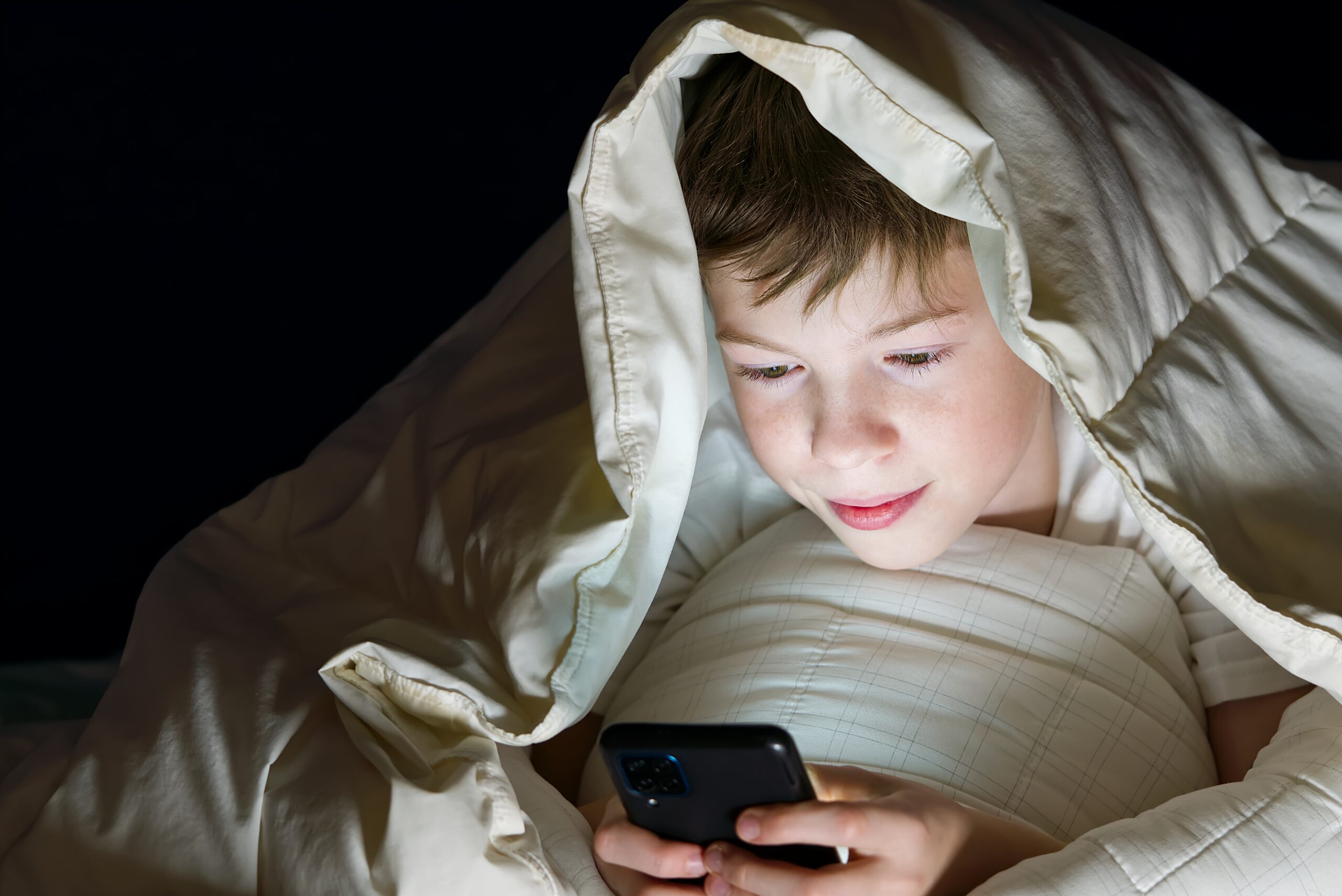 A boy hiding under a blanket while watching a mobile phone late at night, representing the secretive and unhealthy habits linked to smartphone addiction.