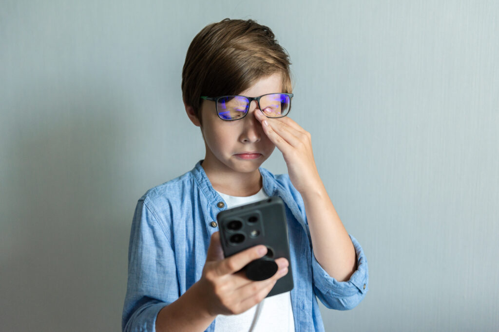 A boy with spectacles on his head rubbing his tired eyes while watching a mobile phone, highlighting the strain and exhaustion caused by smartphone addiction.