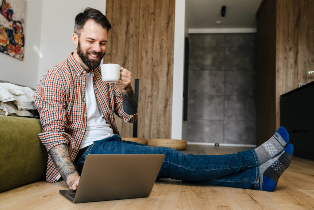 Man working from home on laptop while sitting on the floor with a cup of coffee.