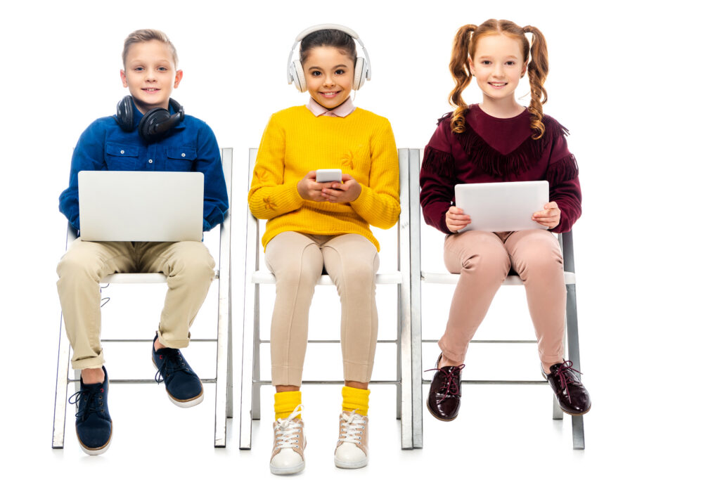 How to learn English – three kids sitting on chairs, one using a tablet, one using a mobile, and another on a laptop while attending online English classes.