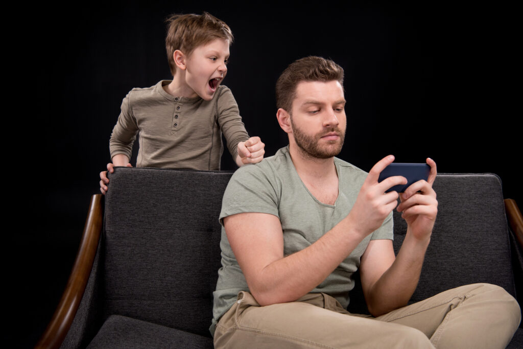 A father calmly using his mobile phone, appearing normal and indifferent, while his child beside him yells for attention. The scene highlights the emotional disconnect and neglect caused by phone addiction effects.