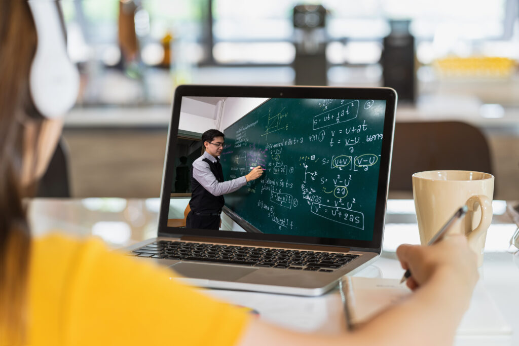 Teacher explaining a lesson through YouTube on a laptop, tablet, and large screen, while students of various ages follow along, representing online learning and digital education.